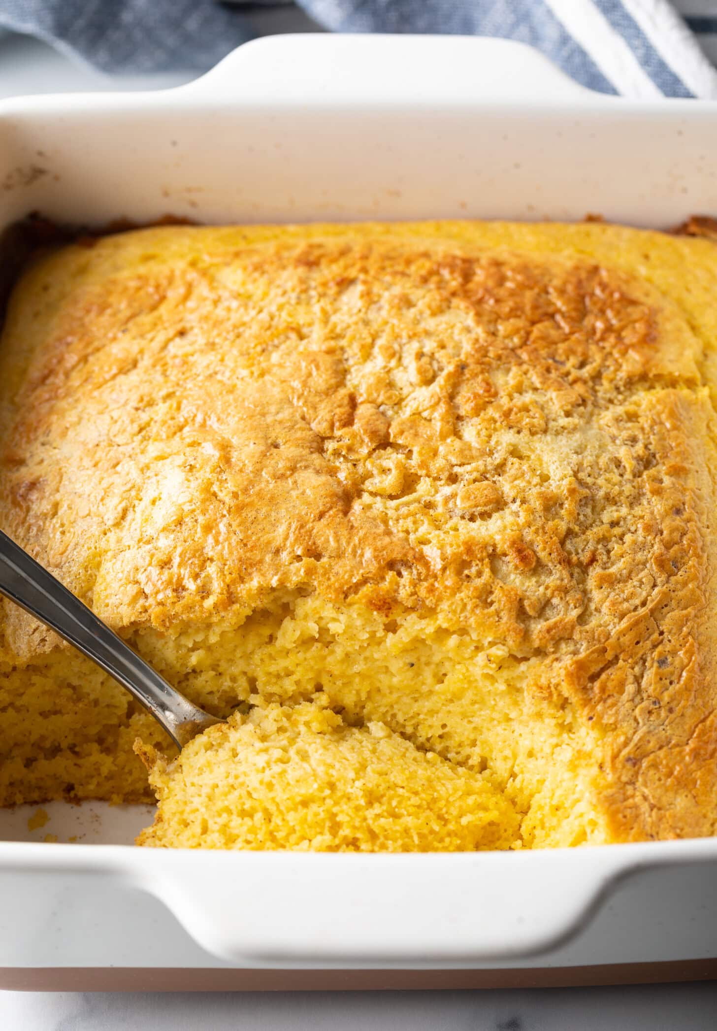 A hand with a metal spoon scooping a portion of corn spoonbread from a square white baking dish.