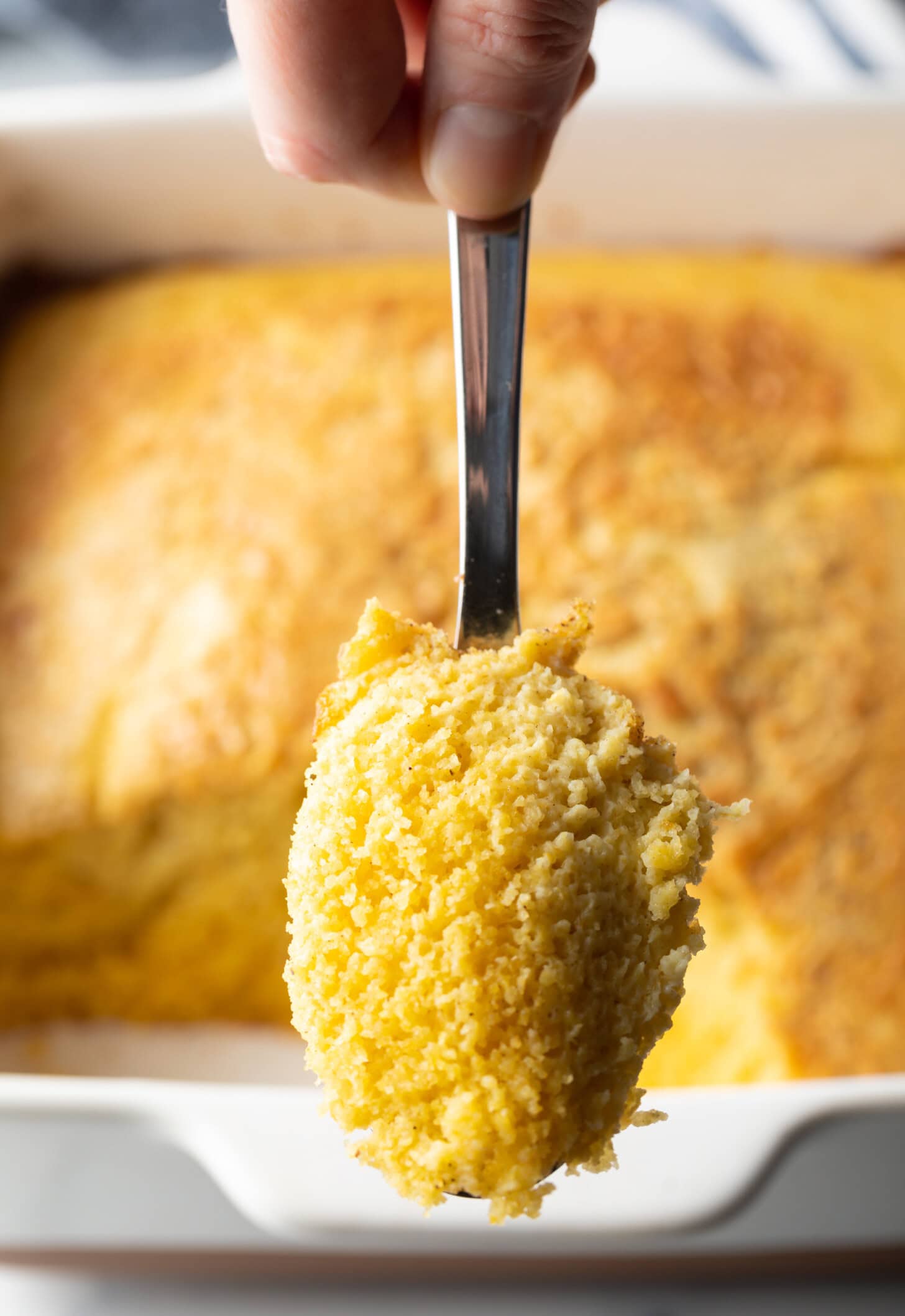A hand holding a metal spoon shows a portion of corn spoon bread to the camera.