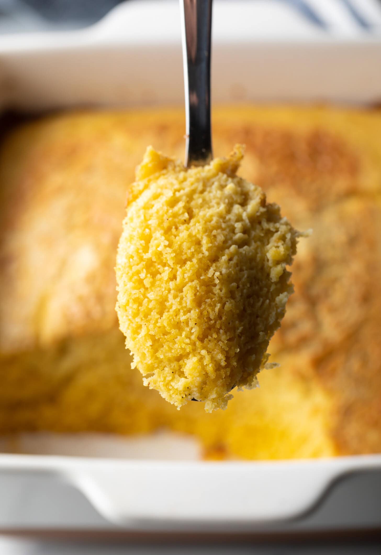 A hand with a metal spoon showing a portion of corn spoon bread to the camera.