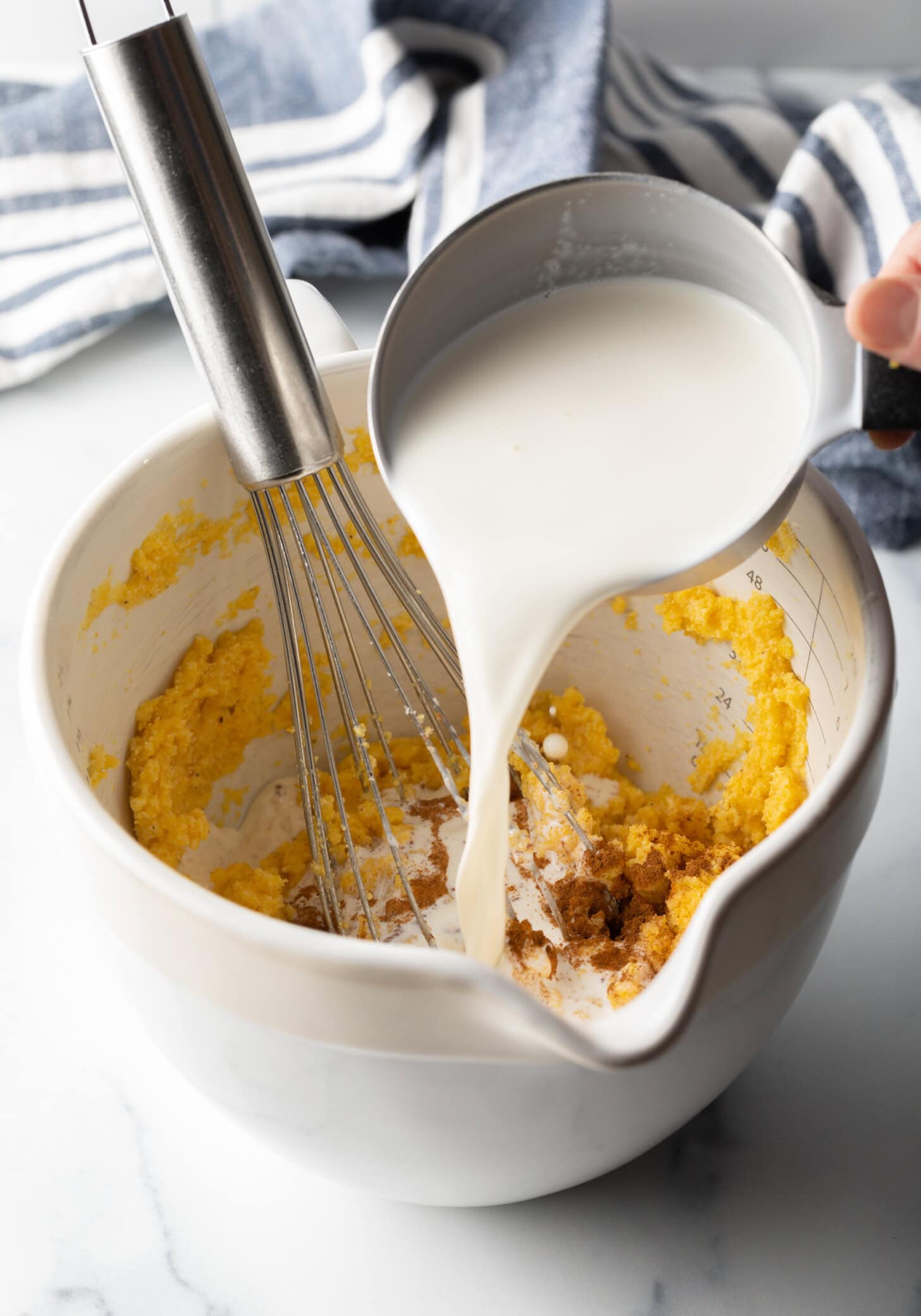 Adding milk to white mixing bowl with spoonbread recipe ingredients and a wire whisk.