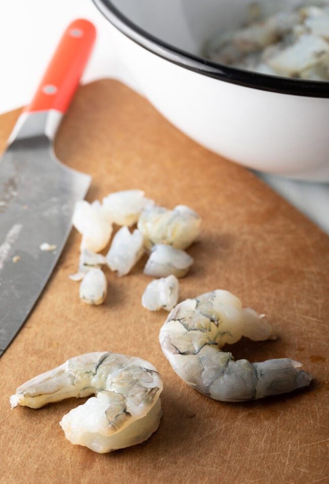 Three fresh peeled and deveined shrimp on a cutting board. One shrimp is chopped into pieces.