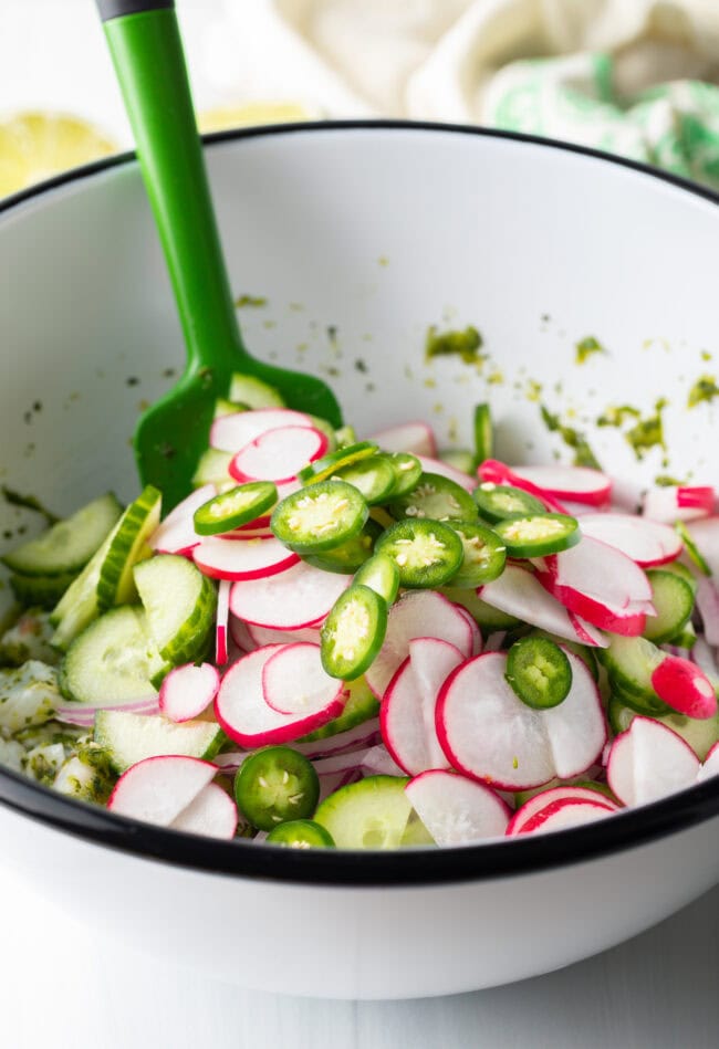 Green spatula in white bowl stirring cucumber, jalapeno, red onions and radishes.
