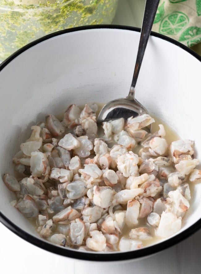 Shrimp pieces marinating in lime juice in a white bowl with metal spoon.