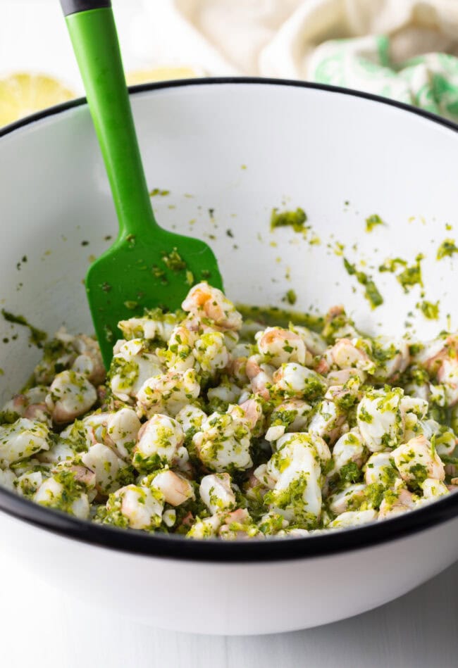 Shrimp combined with cilantro mixture, stirring together with a green spatula in a white bowl.