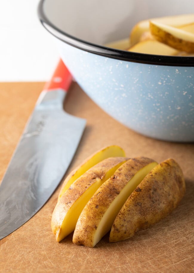 Cutting board and knife, with half a raw potato cut into thick wedges.