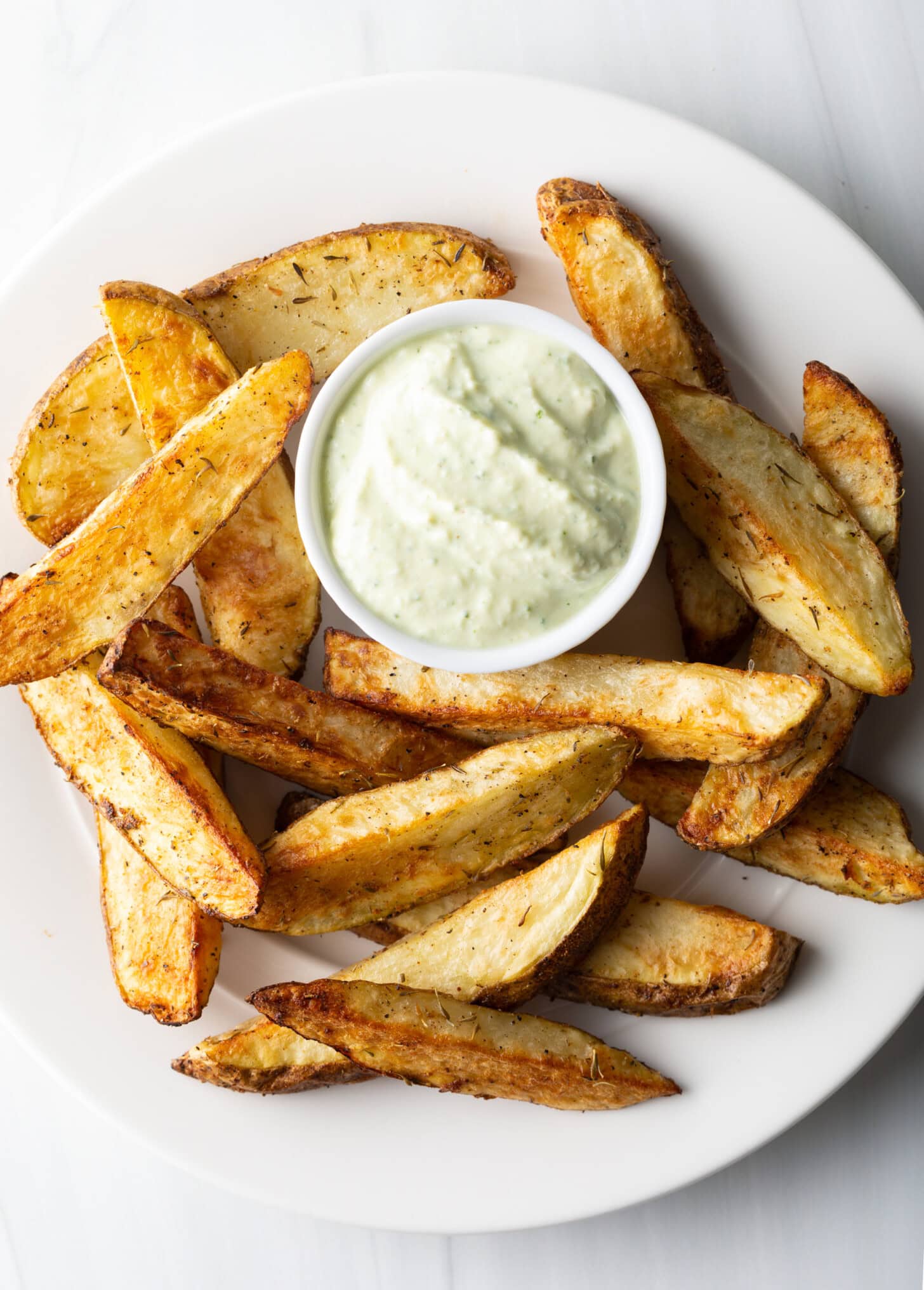 A white plate piled with thick-cut air fryer potato wedges, with a white ramekin of white dipping sauce also on the plate.
