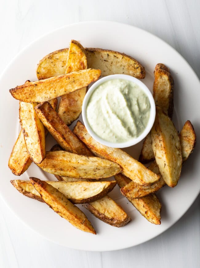 White plate piled with thick cut air fryer potato wedges, with a white ramekin of white dipping sauce also on the plate.