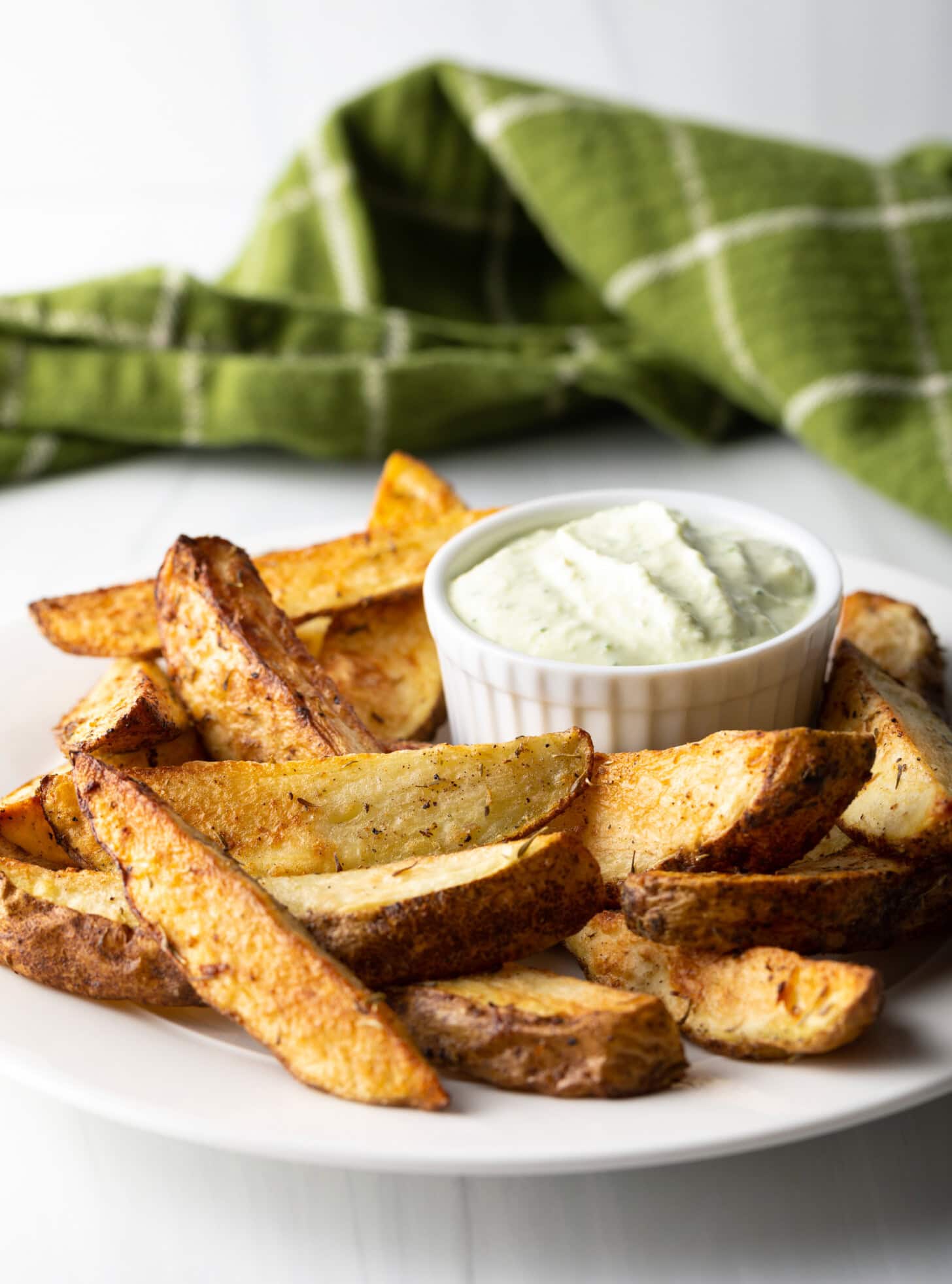 A white plate piled with crispy potato wedges made in the air fryer, with a white ramekin of white dipping sauce also on the plate.