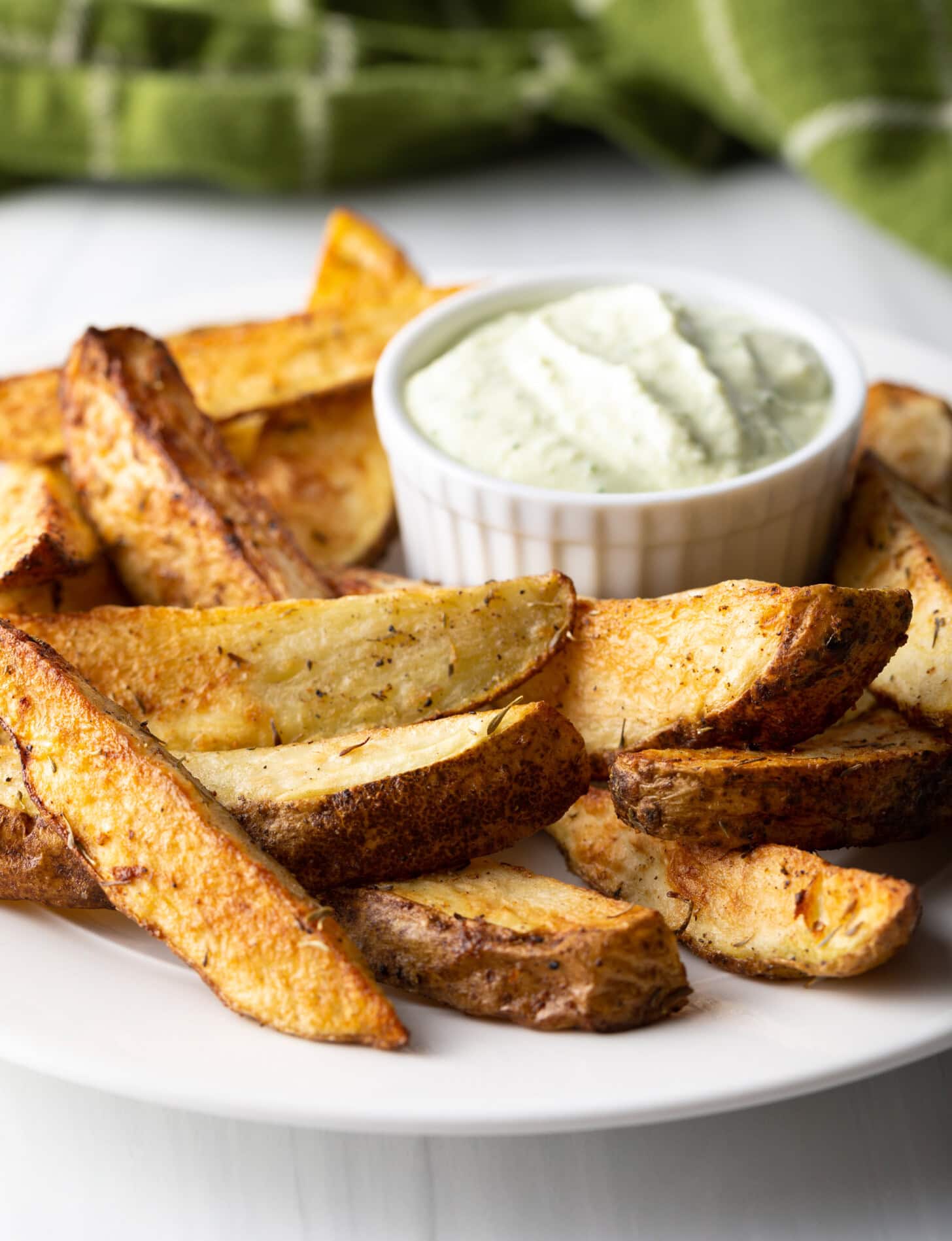 A side view of a plate piled with thick-cut potato wedges made in the air fryer, with a white ramekin of white dipping sauce also on the plate.