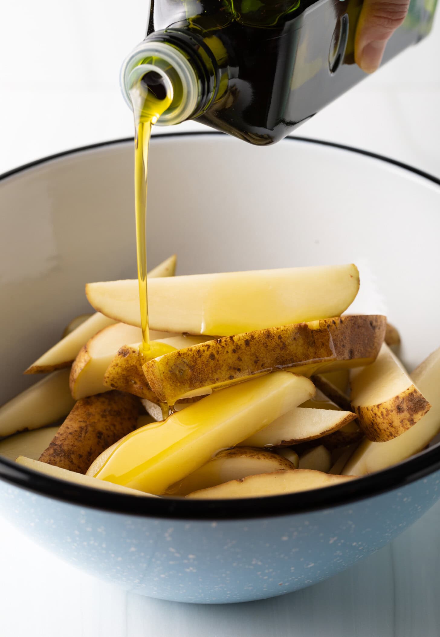 A hand is pouring olive oil onto the air fryer potato wedges in a white bowl.
