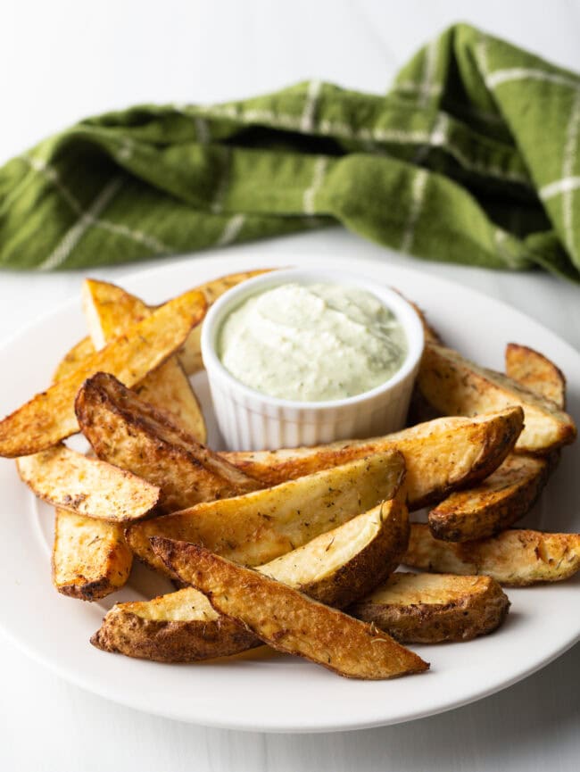 White plate piled with thick cut potato wedges, with a white ramekin of white dipping sauce also on the plate.