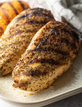 Top down view two whole chicken breasts seasoned and grilled, resting on a white plate.