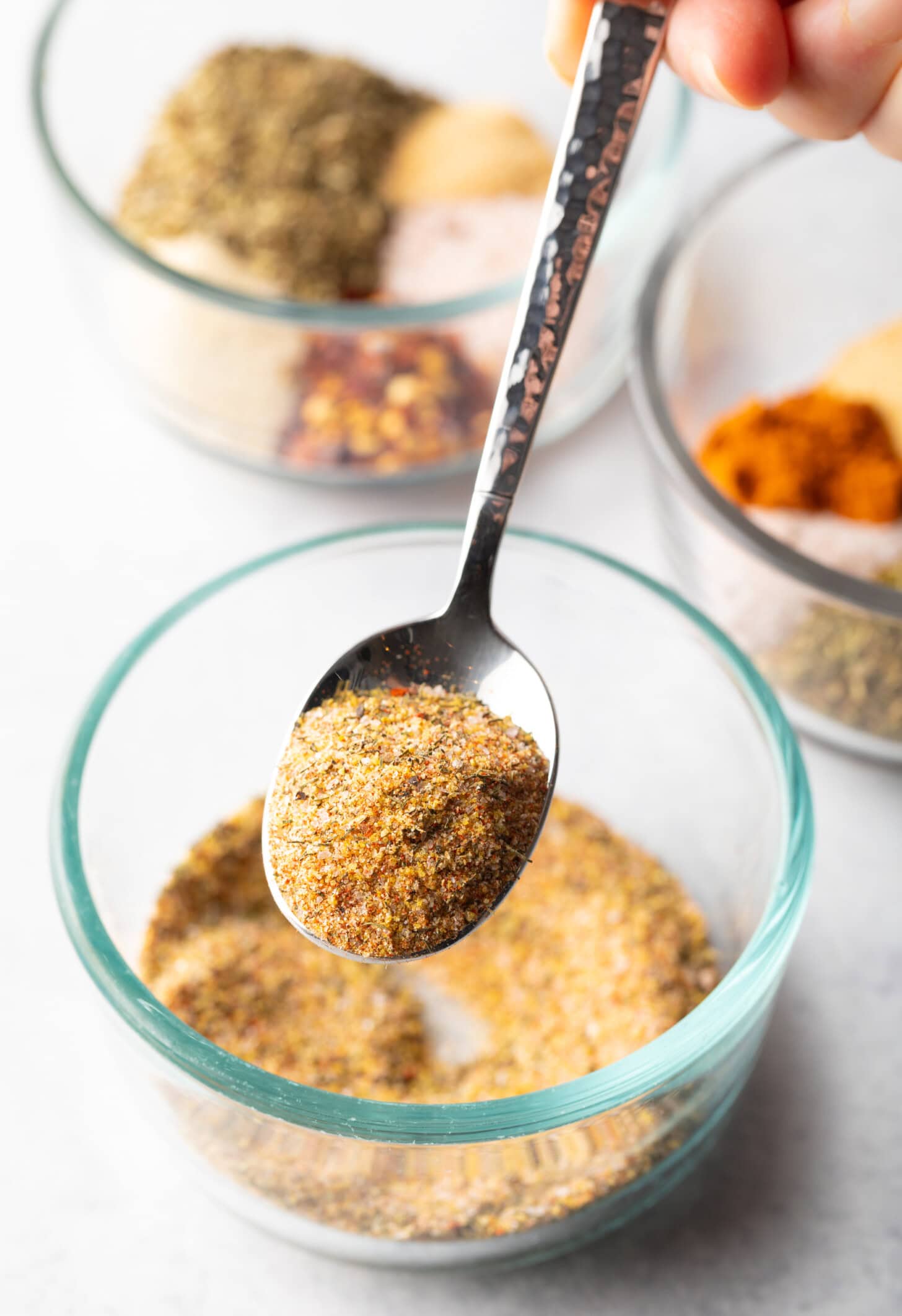 Metal spoon above glass bowl with spice blend. In the spoon is a scoop of seasoning, held to camera.