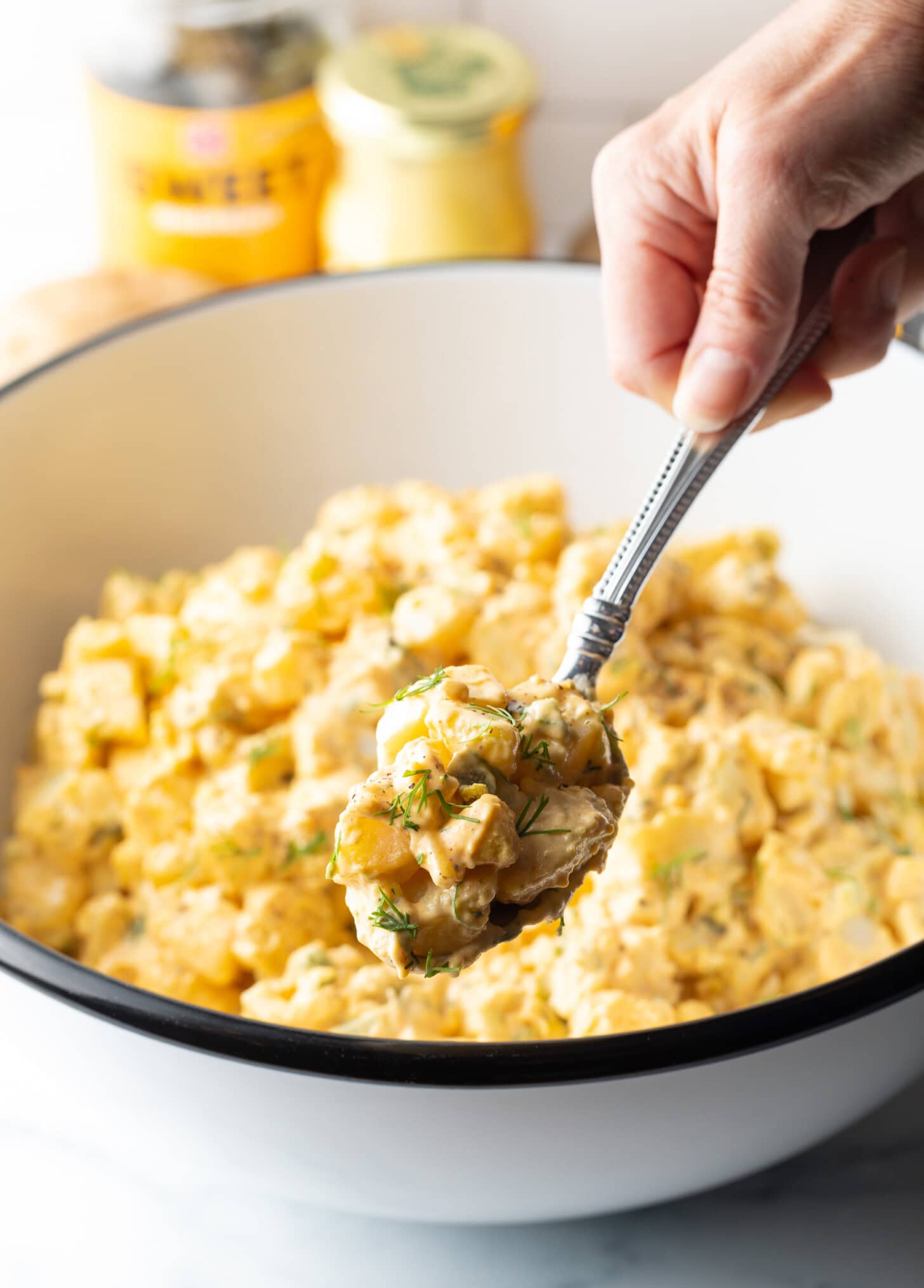 Hand with metal spoon showing a bite of deviled egg potato salad to camera. Jars of pickles and mustard are in the background.