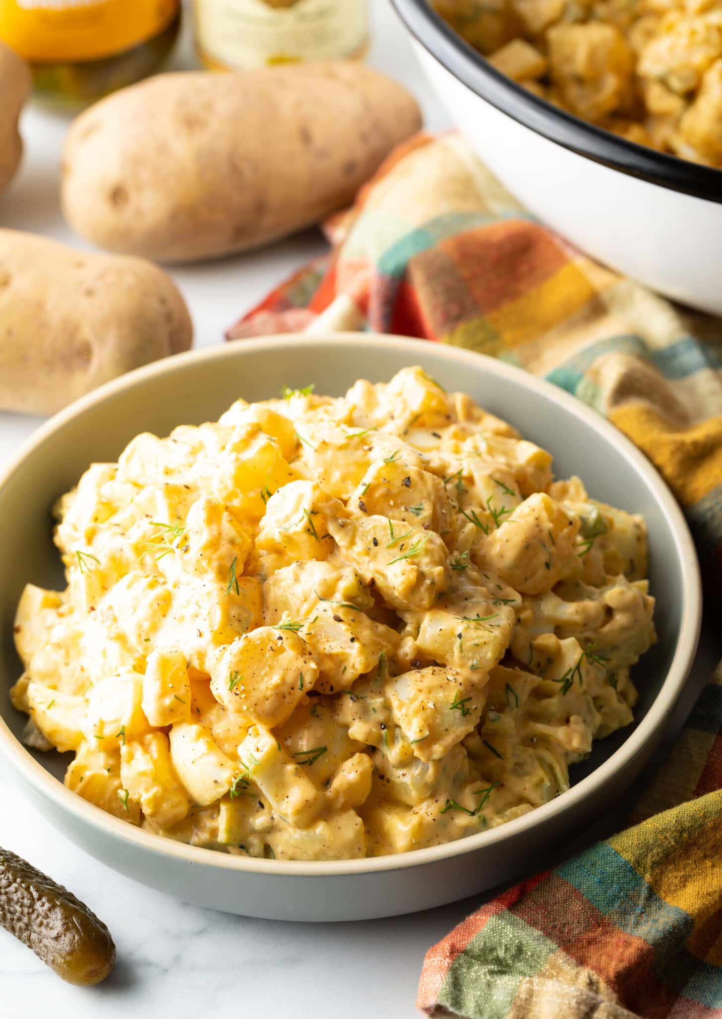 White bowl of potato salad with pickles and potatoes pictured around it.