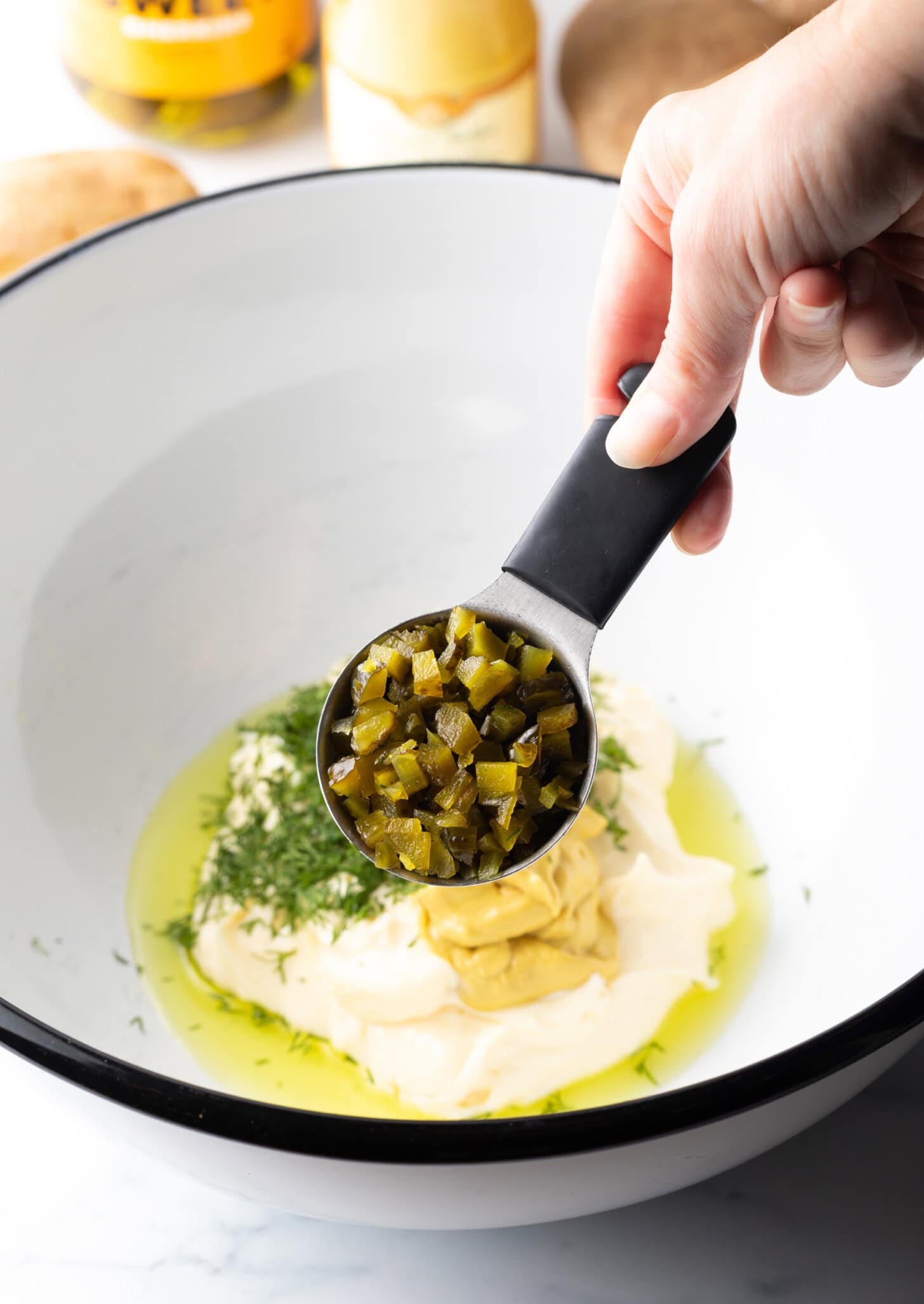 Hand holding measuring spoon with capers, adding to a white mixing bowl with herbs, mayonnaise and mustard in the bottom.