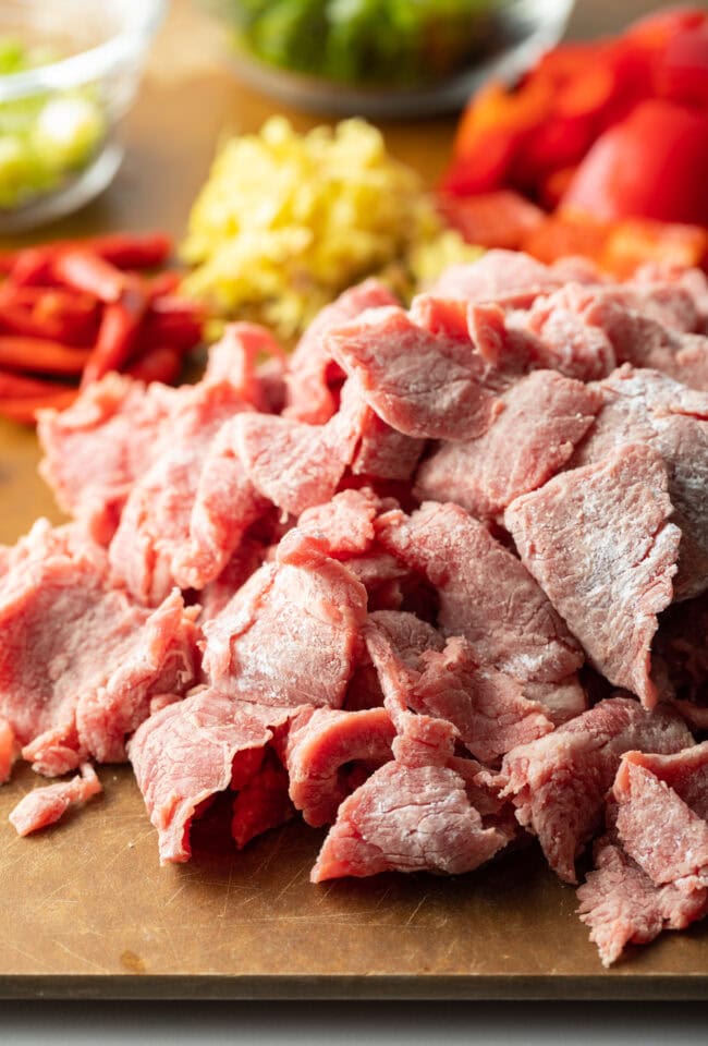 Recipe ingredients chopped on a cutting board in individual piles: steak, onion, red bell peppers, garlic.