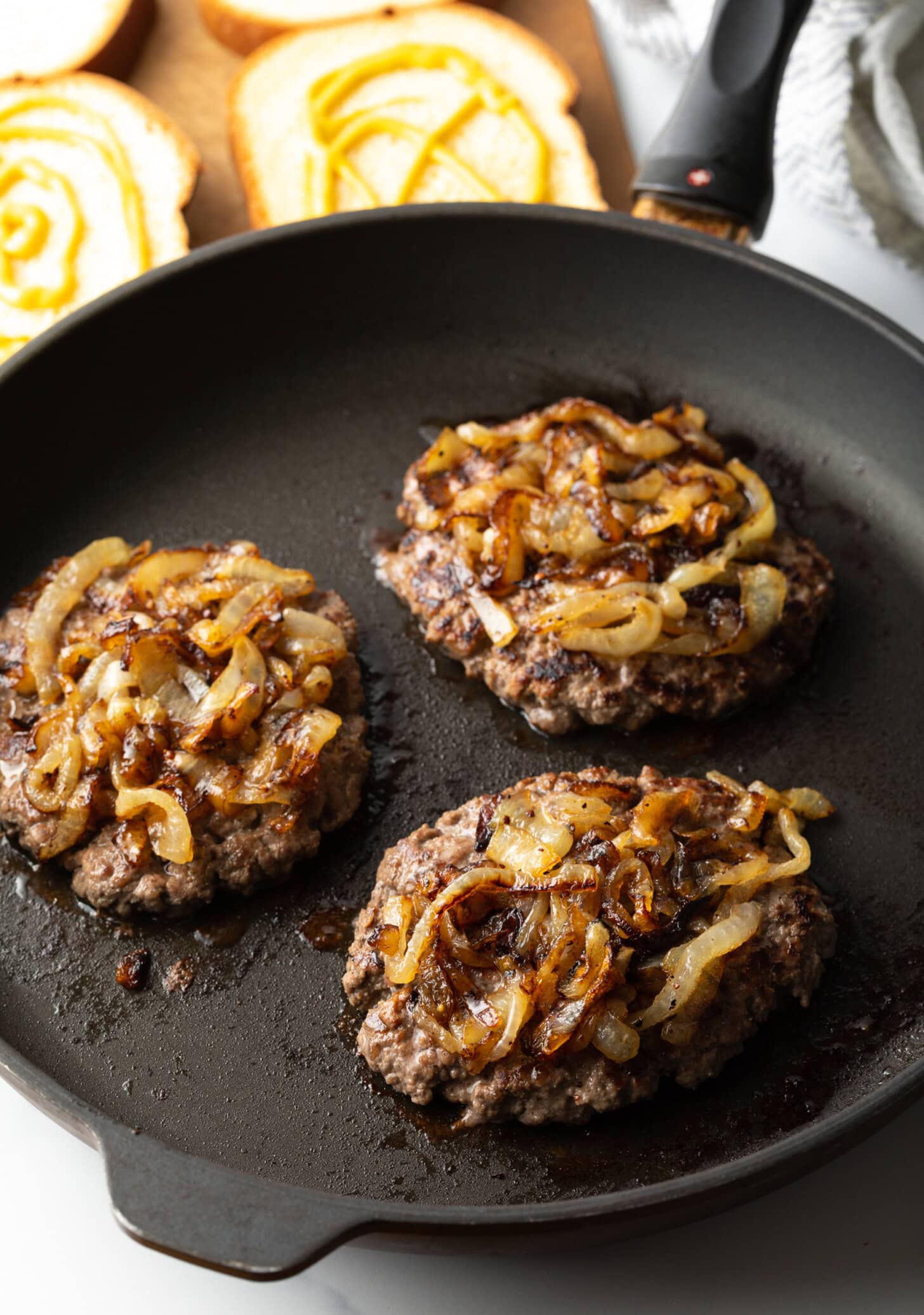 Three burger patties in a skillet, each topped with onion.