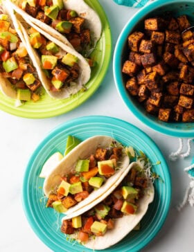 Top down view one green plate and one blue plate each with 3 tacos. Also a blue bowl with more cooked tofu cubes.