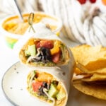 Finished veggie wrap, on a plate with tortilla chips and bowl of hummus in the background.