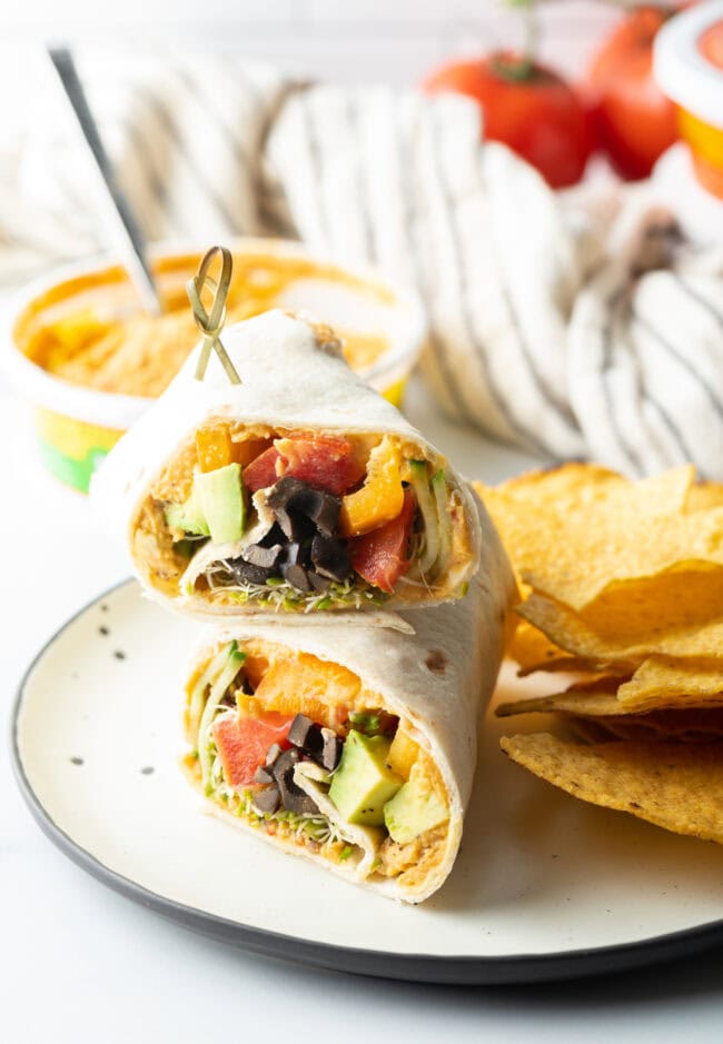 Finished veggie wrap, on a plate with tortilla chips and bowl of hummus in the background.