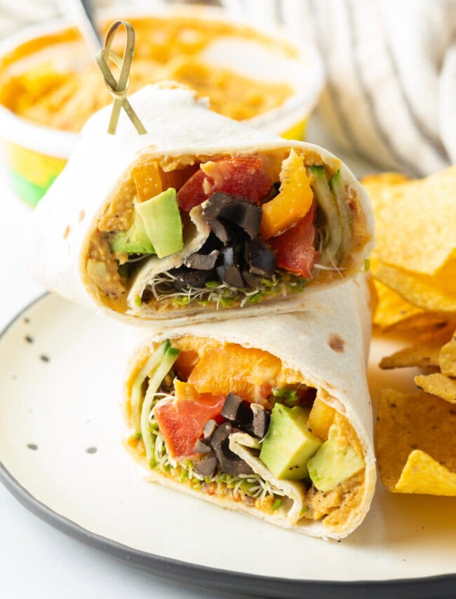 Finished vegetable hummus wrap, on a plate with tortilla chips and bowl of hummus in the background.