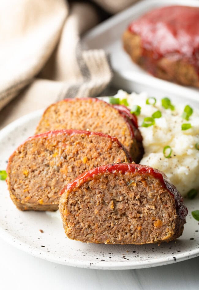 Three slices of air fryer meatloaf on a white plate, with a side of fluffy mashed potatoes and sliced scallions.