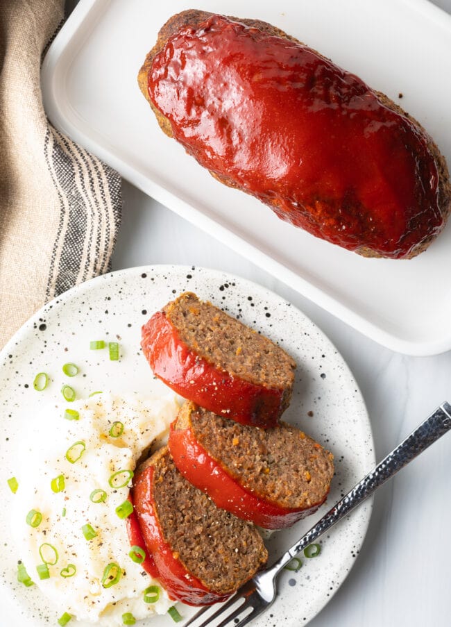 Top down view three slices of meatloaf on a white plate, with a side of fluffy mashed potatoes and sliced scallions.