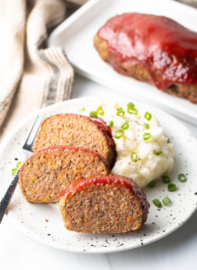 Three slices of meatloaf on a white plate, with a side of fluffy mashed potatoes and sliced scallions.