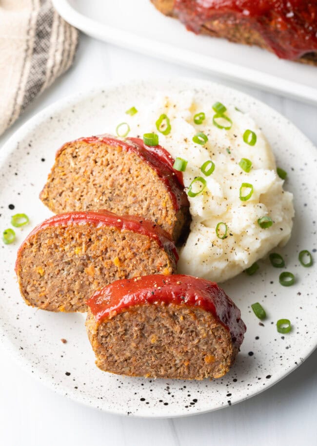 Three slices of meatloaf on a white plate, with a side of fluffy mashed potatoes and sliced scallions.