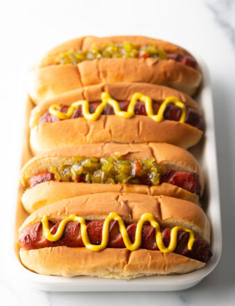 Four hotdogs in buns placed side by side on a white rectangle plate. The toppings alternative between relish and zigzag of mustard.