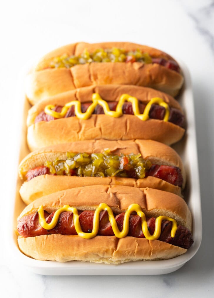 Four hotdogs in buns placed side by side on a white rectangle plate. The toppings alternative between relish and zigzag of mustard.
