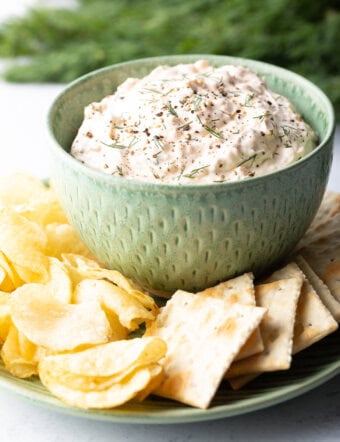 Green bowl filled with creamy clam dip, set on a white plate surrounded by potato chips and crackers.