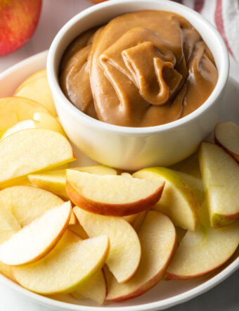 White bowl of cream cheese caramel apple dip, on a white plate surrounded by apple slices.