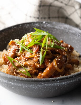 Black bowl of Japanese ginger pork stir fry on top of rice, and garnished with sliced scallions and sesame seeds.