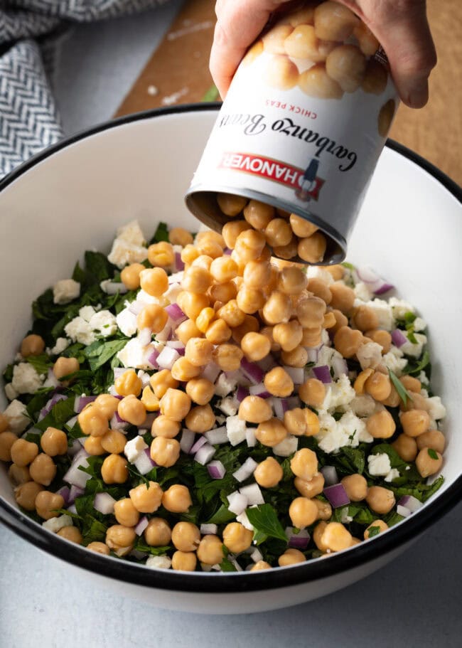 Can of chickpeas being dumped into the large white bowl with other ingredients.