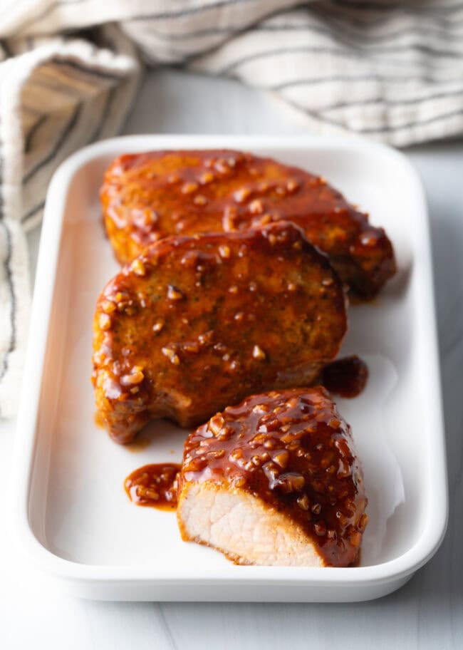 3 pork chops on a rectangle white plate. The chops are all covered with spicy honey garlic soy sauce. One chop has been cut open to show the tender meat inside.