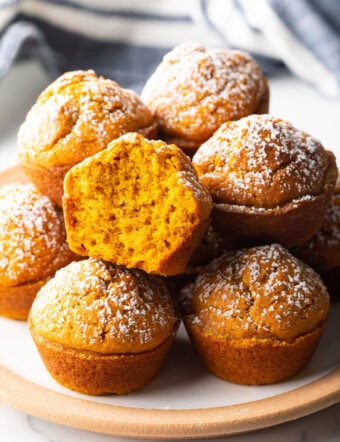 Stack of fluffy muffins made with pumpkin puree, each dusted with powdered sugar. Top muffin has been bitten in half to show inside.
