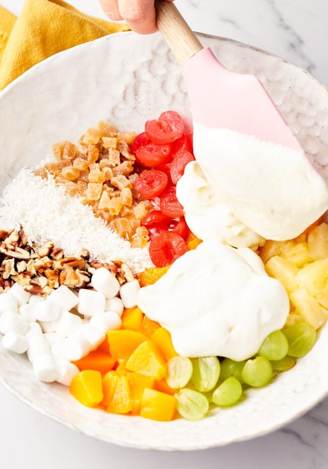 Mixing together canned peaches, maraschino cherries, grapes, pineapple, mini marshmallows, coconut, caramelized ginger, and chopped pecans with cool whip and sour cream.