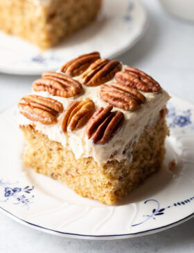 Square of brown butter cake on a plate with pecan halves on top of the white frosted cake.