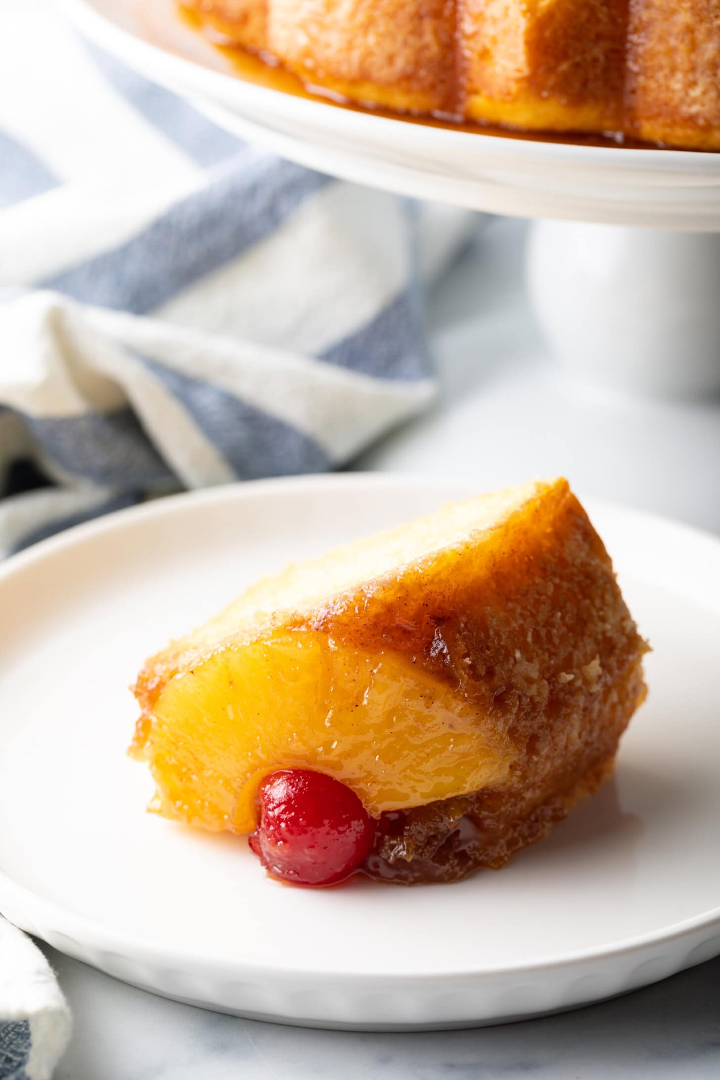 Slice of yellow cake with piece of pineapple and maraschino cherry on a white plate.