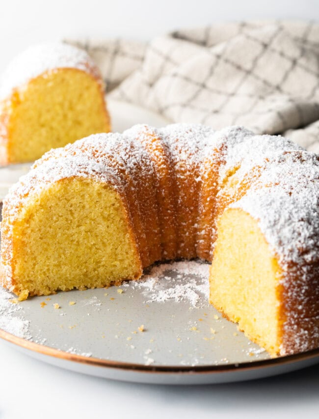 Lemon olive oil bundt cake dusted with powdered sugar on a white plate. A large portion of cake has been removed, showing the inside yellow crumb.
