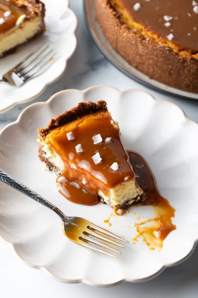 Slice of salted caramel cheesecake on a white plate with a fork. A bite of the cheesecake has been taken out of the tip.