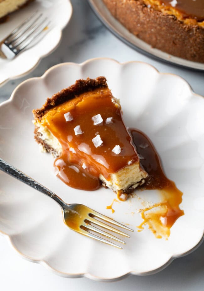 Slice of salted caramel cheesecake on a white plate with a fork. A bite of the cheesecake has been taken out of the tip.