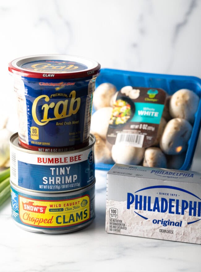 Recipe ingredients: Can of clams, tiny shrimp, and tub of crab stacked on top of each other. A package of cream cheese and package of white mushrooms are next to the stack.