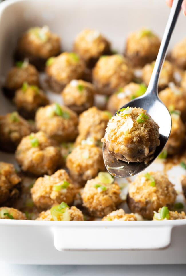 Two dozen seafood stuffed mushrooms in a white baking dish. A metal spoon is showing 1 mushroom to camera.