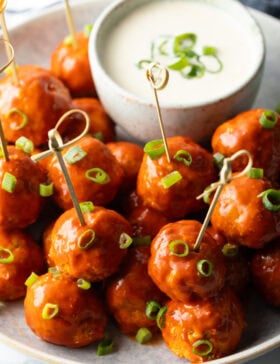 Plate of buffalo chicken meatballs skewered with serving toothpicks, nestled next to a white dish of ranch dressing dipping sauce.