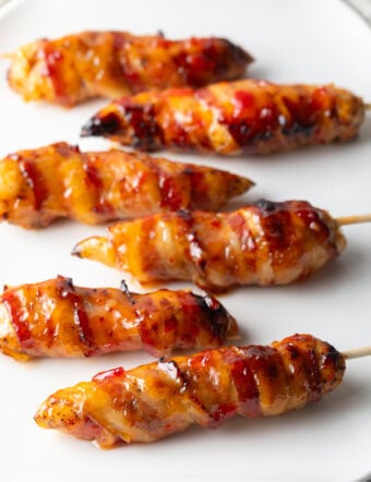 6 skewered bacon wrapped chicken tenders coated with hot sauce on a white plate.