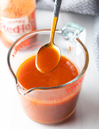 Spoon drizzling buffalo sauce from a large glass measuring cup.