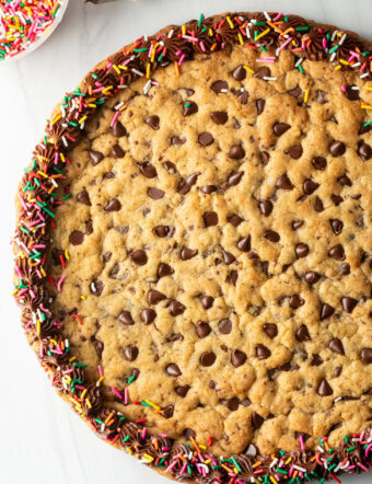 Top down view large chocolate chip cookie cake. The outside edge is piped with chocolate frosting and covered with rainbow sprinkles.